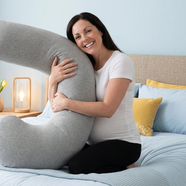 Vrouw omarmt zittend het grijze Ella zwangerschapskussen op bed.